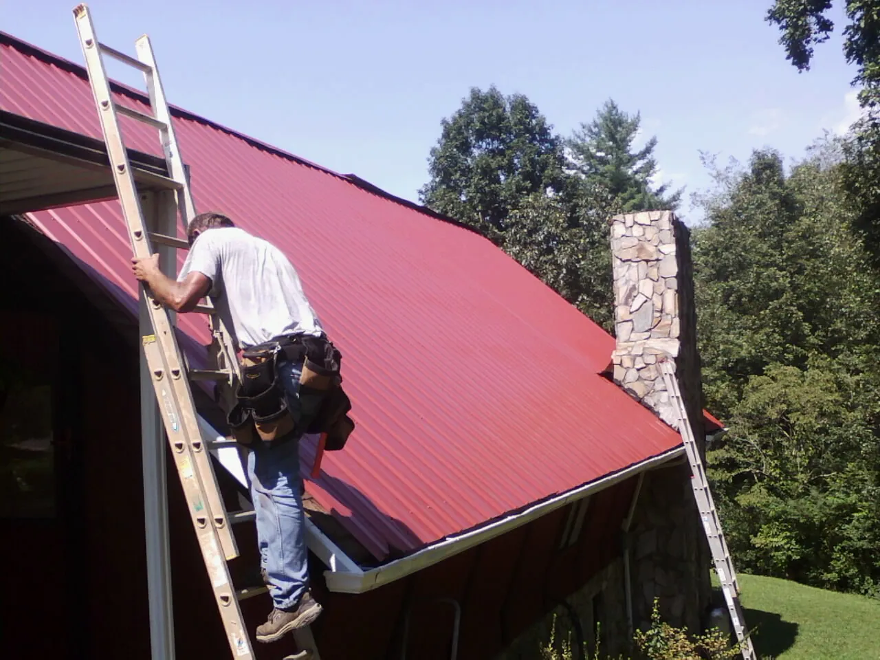 Roofer on a ladder beside a red metal roof and stone chimney — Soffit & Fascia Repair by Roofing Brothers in Auburntown